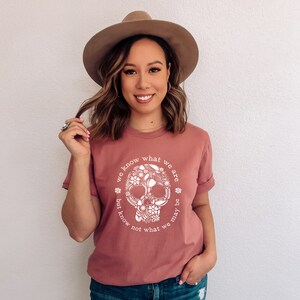 Peut inclure: T-shirt rose poudr&eacute; avec un motif de cr&acirc;ne floral blanc et le texte "we know what we are but know not what we may be". Le t-shirt est &agrave; col rond et a des manches retrouss&eacute;es.