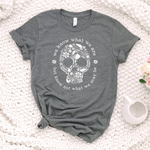 Peut inclure: Un t-shirt gris chin&eacute; avec un motif de cr&acirc;ne floral blanc. Le cr&acirc;ne est entour&eacute; d'un texte circulaire qui dit "we know what we are but know not what we may be". Le t-shirt est en mati&egrave;re douce.