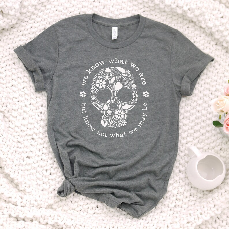 Peut inclure: Un t-shirt gris chin&eacute; avec un motif de cr&acirc;ne floral blanc. Le cr&acirc;ne est entour&eacute; d'un texte circulaire qui dit "we know what we are but know not what we may be". Le t-shirt est en mati&egrave;re douce.