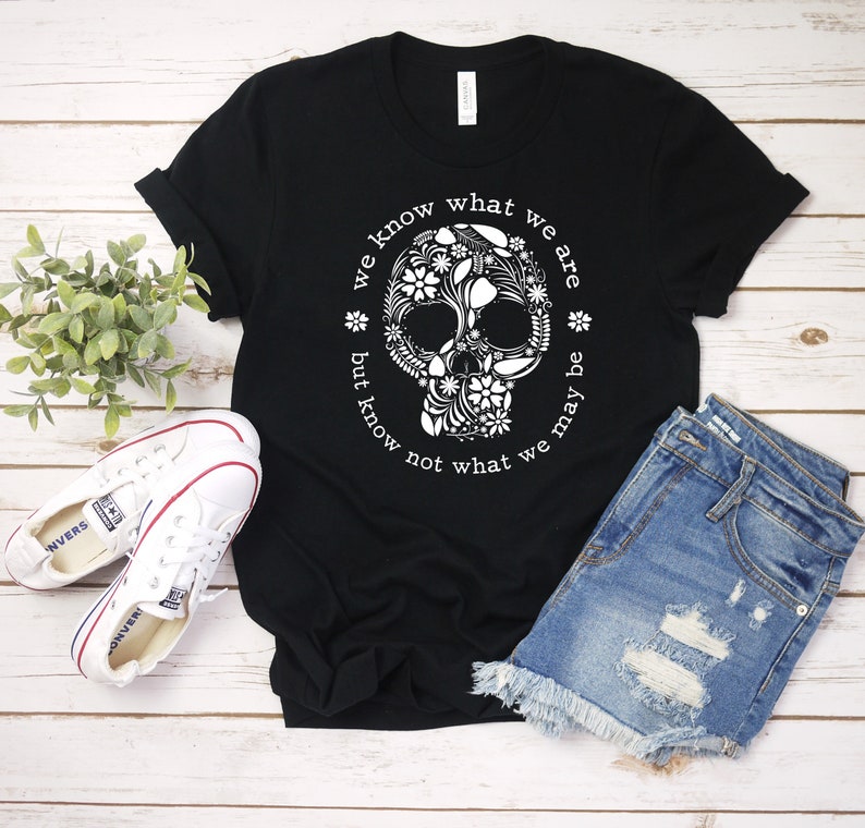 Peut inclure: T-shirt noir avec un motif de cr&acirc;ne floral blanc et le texte "we know what we are but know not what we may be". L'image pr&eacute;sente &eacute;galement un short en jean et des baskets blanches.