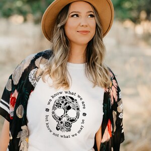 Peut inclure: Une femme porte un chapeau fedora beige et un t-shirt blanc avec un motif de cr&acirc;ne floral noir. Le texte autour du cr&acirc;ne dit "we know what we are but know not what we may be". Elle porte &eacute;galement un ch&acirc;le floral noir.