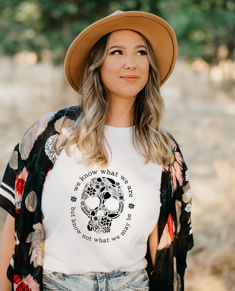 Peut inclure: Une femme porte un chapeau fedora beige et un t-shirt blanc avec un motif de cr&acirc;ne floral noir. Le texte autour du cr&acirc;ne dit "we know what we are but know not what we may be". Elle porte &eacute;galement un ch&acirc;le floral noir.