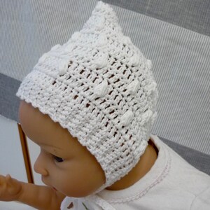 Könnte beinhalten: Eine weiße gehäkelte Babyhaube mit einem strukturierten Muster. Die Haube wird von einem Baby mit hellem Teint getragen.