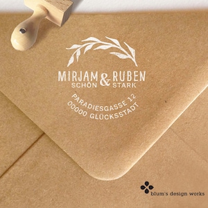 Pode incluir: Um carimbo de madeira com um design floral branco e os nomes "Mirjam & Ruben" impressos em um envelope marrom. O carimbo também inclui o texto "Schön & Stark" e detalhes do endereço.