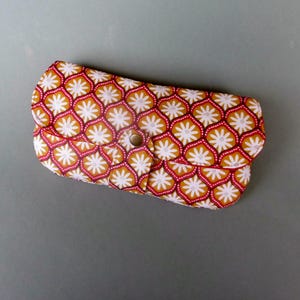 Könnte beinhalten: Ein gemustertes Brillenetui mit Knopfverschluss. Das Etui hat ein sich wiederholendes Blumenmuster in Rot, Gelb und Weiß. Das Etui ist aus einem weichen Material gefertigt und soll Brillen schützen.