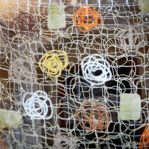 Könnte beinhalten: Ein dekorativer Wandbehang bestehend aus einem cremefarbenen Gitter aus dünner Kordel. Das Gitter ist mit verschiedenen kreisförmigen Garnwirbeln in Orange, Weiß, Gelb und Braun sowie kleinen, quadratischen, hellgrünen Stoffstücken verziert.