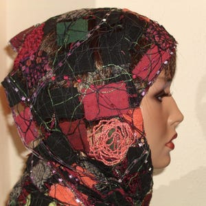 Könnte beinhalten: Eine Kopfbedeckung mit einem Patchwork-Design aus schwarzen, roten, grünen und orangefarbenen Stoffquadraten. Das Stück ist mit einem schwarzen Netz überlagert, das mit dünnen, metallisch schimmernden Fäden und kreisförmigen Stickereien verziert ist.