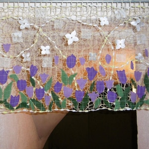 Könnte beinhalten: Ein dekorativer Fensterbehang mit einem gewebten Netzdesign. Er zeigt gestickte lila Tulpen und grüne Blätter am unteren Rand, mit weißen Blumenakzenten. Der Fensterbehang hat eine hellbeige Farbe. Die Maße sind nicht sichtbar.