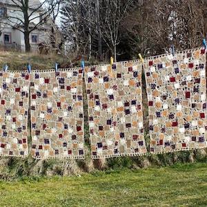 Könnte beinhalten: Mehrere handgefertigte Häkeldecken hängen an einer Wäscheleine. Die Decken weisen ein sich wiederholendes Muster aus Quadraten in Beige, Braun, Burgunderrot und Lila auf. Die Decken sind mit farbigen Wäscheklammern befestigt und trocknen in der Sonne.
