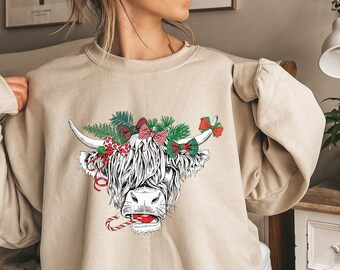 May include: A beige sweatshirt with a Christmas-themed illustration of a Highland cow wearing a wreath of greenery and red bows. The cow is holding a candy cane in its mouth. The text "Sand" is printed on the bottom left corner of the sweatshirt.