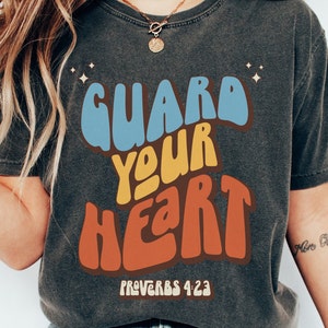 May include: A dark gray t-shirt with a retro-style graphic that says "GUARD YOUR HEART" in colorful letters. The text "PROVERBS 4:23" is printed below the graphic.