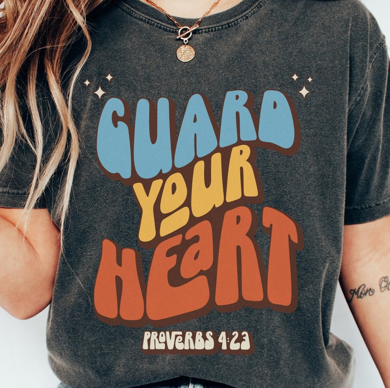 May include: A dark gray t-shirt with a retro-style graphic that says "GUARD YOUR HEART" in colorful letters. The text "PROVERBS 4:23" is printed below the graphic.