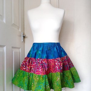 May include: A colourful tiered skirt on a mannequin, featuring a batik-style pattern. The skirt has a blue elasticated waistband and three layers in blue, pink, and green. The skirt's design includes three distinct layers, each with a unique colour and pattern.