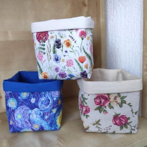 May include: Three fabric storage baskets with different floral patterns. The baskets are made of a soft, natural fabric and have a white lining. The patterns include a white background with colourful flowers, a white background with pink roses, and a blue background with a swirling pattern.