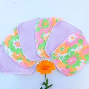 May include: Set of six reusable makeup remover pads. The pads are made of soft, absorbent fabric and have a floral print in shades of pink, green, and orange. The pads are arranged in a fan shape and are surrounded by a single orange flower.