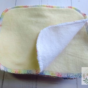 May include: A yellow cloth burp cloth with a white terry cloth side. The cloth has a rainbow colored stitched border.