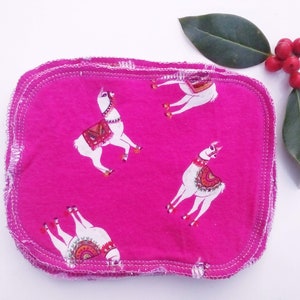 May include: A hot pink, square-shaped cloth with a white stitched border. The cloth is printed with a pattern of white llamas wearing colorful blankets. A sprig of red berries and green leaves are in the background.