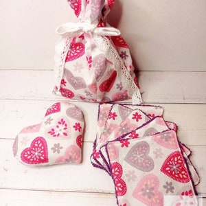 May include: A set of pink and white heart-shaped fabric squares with a purple trim. The squares are stacked on top of each other. There is a pink and white heart-shaped fabric bag with a white lace trim and a white ribbon tied around it. The bag is sitting on a white wooden surface.