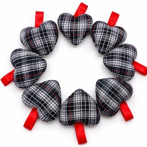 May include: A circular arrangement of heart-shaped fabric ornaments. Each heart is patterned with a black, white, and red plaid design and has a bright red ribbon attached. The hearts are arranged against a white background.