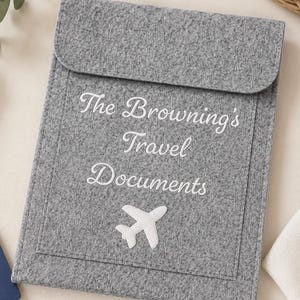 May include: A gray felt travel document holder with a flap closure. The holder features the embroidered text "The Browning's Travel Documents" and a white airplane applique. The holder is designed to store travel documents.