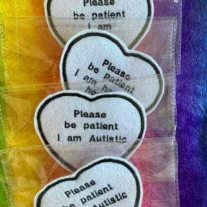 Puede incluir: Cuatro parches de fieltro blanco en forma de corazón con texto bordado en negro. El texto en los parches dice: "Please be patient I am", "Please be patient I am he", "Please be patient I am Autistic", y "Please be patient I am Autistic".