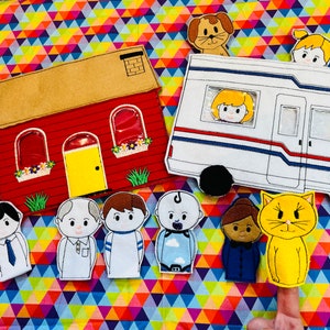 May include: A set of felt finger puppets featuring a red house, a white camper van, a yellow cat, a baby, a girl with brown hair, a boy with brown hair, a man with white hair, and a girl with black hair. The puppets are on a colorful background.