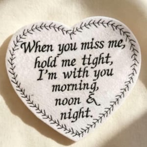 May include: A white heart-shaped felt ornament featuring a black stitched border and the black printed text: When you miss me, hold me tight, I'm with you morning, noon & night. The object has a soft, textured surface and a simple, clean design.