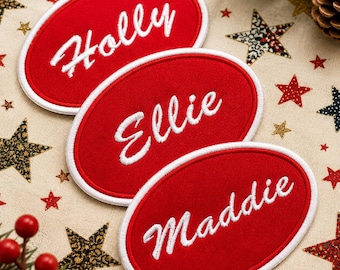 Puede incluir: Tres etiquetas ovaladas rojas con nombres bordados en blanco: Holly, Ellie y Maddie. Cada etiqueta tiene un borde blanco. Están sobre un fondo festivo con decoraciones de estrellas y bayas.