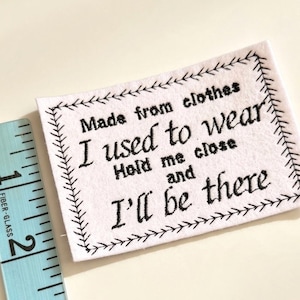 May include: A rectangular white fabric patch with black embroidered text reading Made from clothes I used to wear Hold me close and I'll be there, framed by a stitched border, next to a light blue measuring tape showing 5 centimetres.