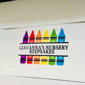 May include: A white fabric envelope with a rectangular design featuring a row of colourful crayons. The text "Giavanna's Nursery Keepsakes" is printed in black. The crayons are in shades of purple, red, orange, yellow, green, and blue.