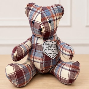 May include: A handmade teddy bear with a plaid pattern in shades of burgundy, cream, and blue. The bear has a heart-shaped patch with text that reads "Made from...hug me tight...Love, Great dana!" The bear is sitting on a wooden surface.