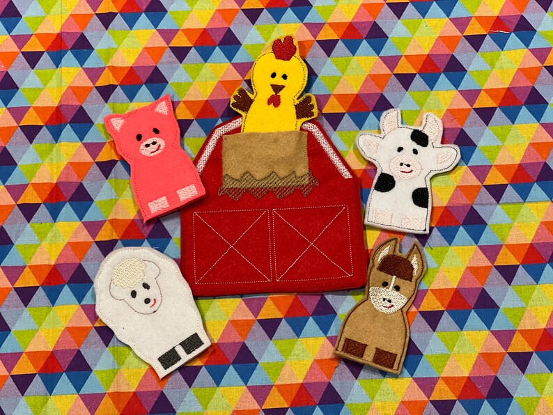 Peut inclure: Une grange en feutre rouge avec un toit marron, entour&eacute;e de cinq marionnettes &agrave; doigts : un cochon rose, un poulet jaune, une vache blanche, un mouton blanc et un &acirc;ne marron. Le fond est un motif g&eacute;om&eacute;trique color&eacute;.