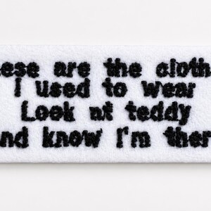Könnte beinhalten: Ein rechteckiger, weißer Stofffleck mit schwarzem, gesticktem Text. Der Text lautet: "Das sind die Kleider, die ich trug. Schau Teddy an und wisse, dass ich da bin."
