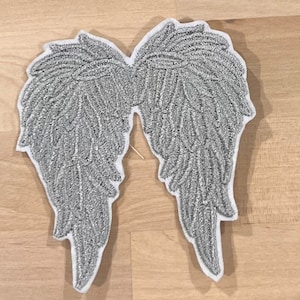 Può includere: Un paio di ali d'angelo in glitter argentato con un retro in feltro bianco. Le ali sono ricamate con un motivo a piume dettagliato.