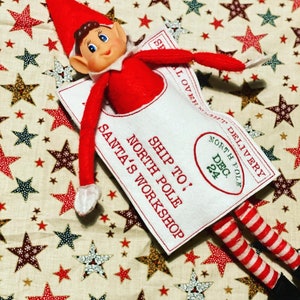 May include: A red and white elf doll wearing a white fabric envelope with the text "Ship to: North Pole Santa's Workshop" and a red circle with the text "North Pole Dec. 24"