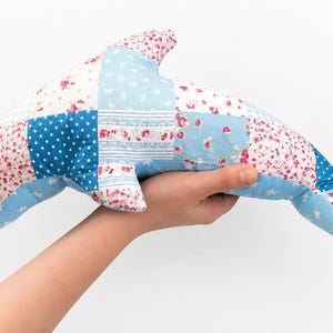 May include: A patchwork dolphin-shaped pillow in shades of blue, white, and pink. The pillow is made of fabric squares with floral, polka dot, and star patterns. The dolphin is held in a hand against a white background.