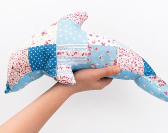 May include: A patchwork dolphin-shaped pillow in shades of blue, white, and pink. The pillow is made of fabric squares with floral, polka dot, and star patterns. The dolphin is held in a hand against a white background.
