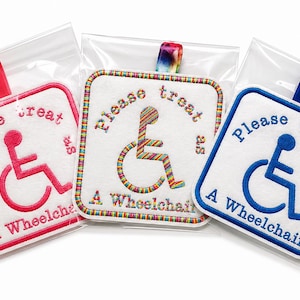 Op de afbeelding: Drie geborduurde patches in doorzichtige plastic zakken. Elke patch heeft een rolstoelsymbool en de tekst "Please treat as A Wheelchair" op een witte achtergrond. De patches zijn in rood, regenboog en blauw.