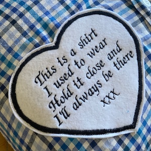 May include: White heart-shaped patch with black stitching around the edges. The patch has black embroidered text that reads "This is a shirt I used to wear Hold it close and I'll always be there XXX".