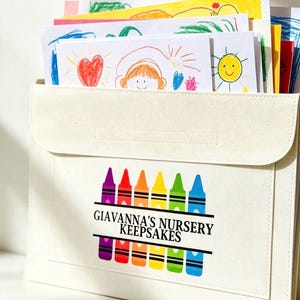 May include: A cream-colored fabric document organizer with a flap closure, featuring a graphic of colorful crayons and the black text GIAVANNA'S NURSERY KEEPSAKES. The organizer is filled with various white papers containing colorful children's drawings.