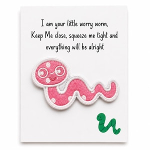 Könnte beinhalten: Eine weiße Karte mit dem Text "I am your little worry worm, Keep Me close, squeeze me tight and everything will be alright." Ein rosa Wurmaufnäher mit weißen Punkten und Brille ist abgebildet, zusammen mit einem kleinen grünen Wurm.