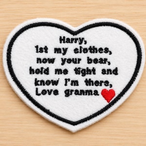 May include: A white heart-shaped patch with black embroidered trim and text. The text reads "Harry, 1st my clothes, now your bear, hold me tight and know I'm there, Love granma" with a small red heart.