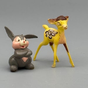May include: A small gray rabbit figurine with a pink face and chest sitting with arms crossed, positioned next to a standing yellow fawn figurine with brown head markings and white spots on its back, both featuring painted facial details.