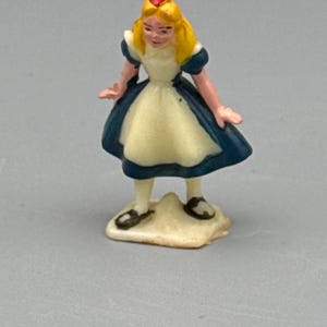 May include: A small figurine of a character with blonde hair, a red headband, a blue dress with short puff sleeves, and a white apron. The figure stands on a small, irregular off-white base and wears dark shoes with white socks.