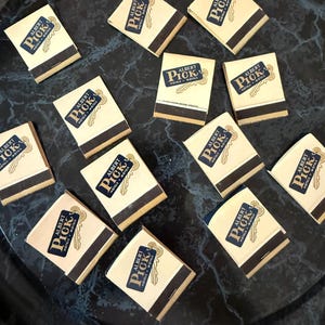 May include: A collection of vintage matchbooks with the "Albert Pick Hotels & Motels" logo. The matchbooks are cream-colored with a black stripe and a navy blue and gold logo. They are arranged on a dark, marbled surface.