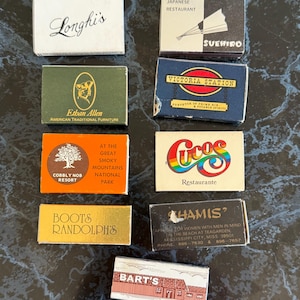 May include: A collection of vintage matchboxes with various designs and text. The matchboxes feature logos and names such as "Longhi's", "Suehiro", "Victoria Station", "Cocos Restaurante", and "Khamis". The matchboxes are arranged on a dark, textured surface.