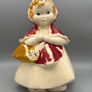 May include: A ceramic figurine of a child wearing a red hooded cape and a cream-colored dress. The figure has blonde hair, blue eyes, and holds a yellow basket with a small green floral design. The red paint on the cape shows signs of wear.
