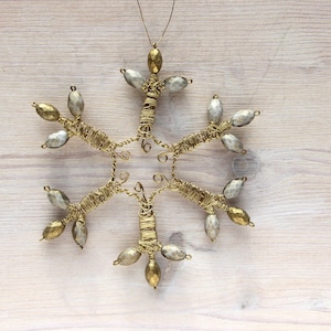 Könnte beinhalten: Handgefertigter Schneeflocken-Schmuck aus goldfarbenem Draht und silbernen und goldfarbenen Perlen. Der sechszackige Stern hat eine Schlaufe zum Aufhängen. Die Dekoration liegt auf hellem Holz. Eine festliche Weihnachtsdekoration.