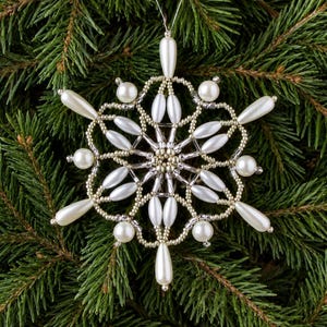 Könnte beinhalten: Ein Schneeflocken-Ornament aus weißen tropfenförmigen Perlen, runden weißen Perlen und kleinen metallisch silbernen Saatperlen. Das filigrane Design zeigt ein symmetrisches Sternmuster mit einem zentralen Cluster aus Silberperlen.