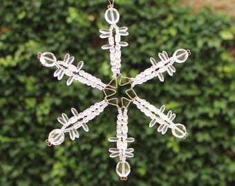 Pode incluir: Um enfeite de floco de neve transparente com contas e uma estrutura de arame dourado. O floco de neve tem seis pontas, cada uma com uma série de contas transparentes e grupos de contas decorativas menores. Pendurado por um fio dourado fino.